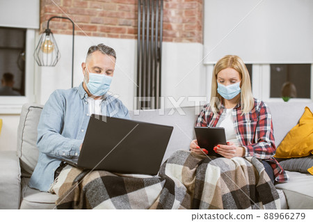 Mature couple in face masks using laptop and tablet on couch 88966279
