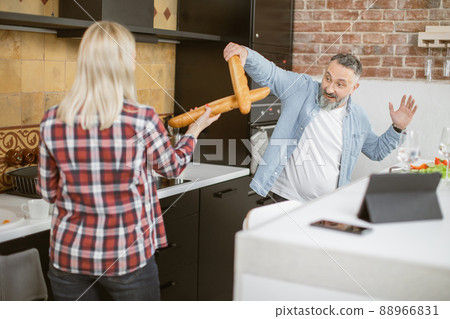 Joyful mature couple fighting with baguettes on kitchen Joyful mature couple fighting with baguettes on kitchen 88966831