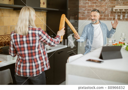 Joyful mature couple fighting with baguettes on kitchen Joyful mature couple fighting with baguettes on kitchen 88966832