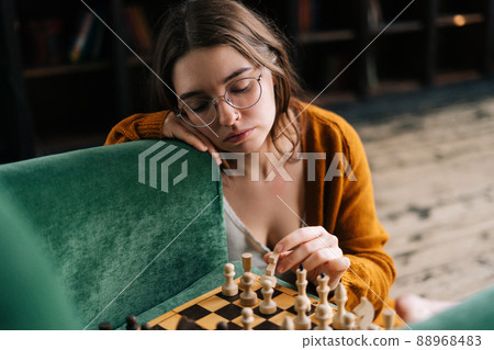 Close-up of pensive cute young woman wearing elegant eyeglasses making chess move with knight piece sitting in wooden floor. Pretty intelligent lady playing logical board game alone at home. Close-up of pensive cute young woman wearing elegant eyeglasses making chess move with knight piece sitting in wooden floor. Pretty intelligent lady playing logical board game alone at home. 88968483