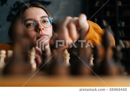 Close-up low-angle view of young woman in elegant eyeglasses making chess move with knight piece sitting in floor, looking at camera. Pretty intelligent lady playing logical board game alone at home. Close-up low-angle view of young woman in elegant eyeglasses making chess move with knight piece sitting in floor, looking at camera. Pretty intelligent lady playing logical board game alone at home. 88968488