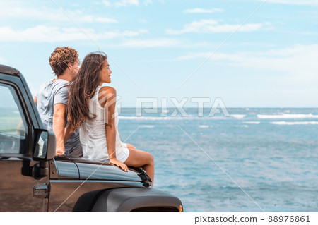 Car road trip travel couple tourists enjoying ocean view relaxing on hood of sports utility car. Happy Asian woman, man friends smiling on beach. 88976861