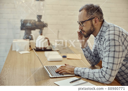 Bearded man talking on cellphone and using laptop in cafe Bearded man talking on cellphone and using laptop in cafe 88980771