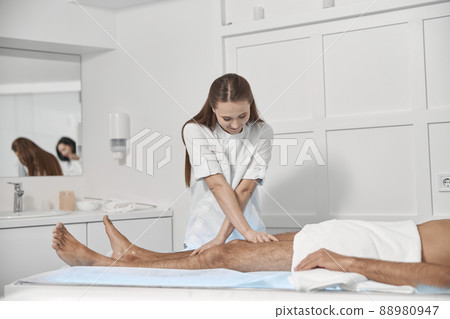 Lady in white uniform massages legs of middle aged man in contemporary clinic Lady in white uniform massages legs of middle aged man in contemporary clinic 88980947