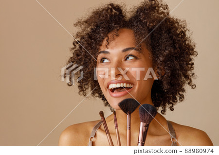 Cheerful woman demonstrating makeup tools against beige wall 88980974