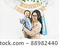 Asian baby boy celebrating first birthday,A mother sits and hug her son smiling on a floor with a minimal background,white balloons and birthday flag.Selective focus 88982440