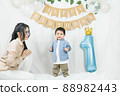 Asian baby boy celebrating first birthday,A mother sits and looks at her son standing proudly on a floor with a minimal background,white balloons and birthday flag.Selective focus 88982443