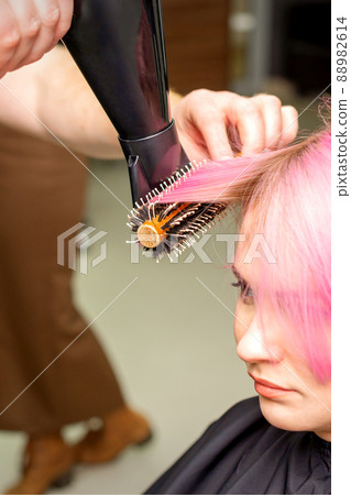 Drying short pink hair of young caucasian woman with a black hairdryer and black round brush by hands of a male hairdresser in a hair salon, close up 88982614