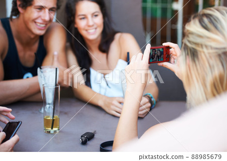Capturing their happiest memories. Group of teens enjoying beverages while at an outdoor restaurant while taking a photo with a mobile phone. Capturing their happiest memories. Group of teens enjoying beverages while at an outdoor restaurant while taking a photo with a mobile phone. 88985679
