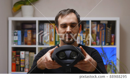 A Caucasian man in a black hoodie holding a virtual reality helmet. Home interior. VR glasses for entertainment, learning and gaming. 88988295