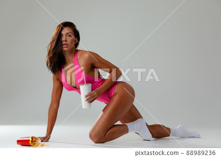 cheat meal concept. athlete woman is posing with fast food on light gray background, studio shot. young sporty woman is holding fast food close up. cheat meal concept. athlete woman is posing with fast food on light gray background, studio shot. young sporty woman is holding fast food close up. 88989236