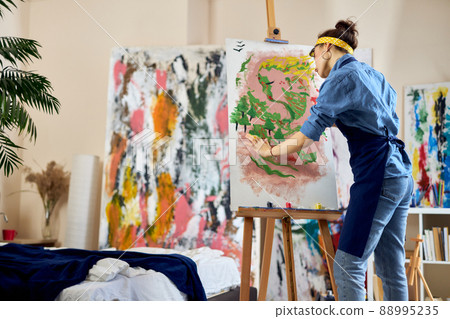 Creative woman, female artist in apron looking inspired while creating her own abstract painting at home studio workshop 88995235