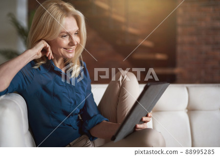 Smiling mature woman watching video using digital tablet pc, relaxing on a couch at home in the living room 88995285