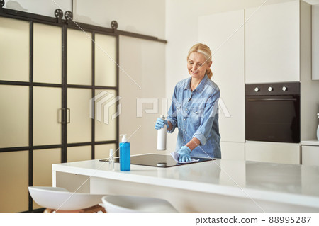 Smiling mature housewife in rubber gloves looking busy while cleaning kitchen surface using cloth and spray detergent at home in the daytime 88995287