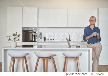 Content mature businesswoman in glasses smiling at camera, holding a cup of coffee and using laptop, standing in modern kitchen 88995288