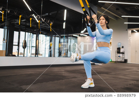 Athletic woman doing squat exercise, training legs and glutes muscular with fitness straps. TRX exercise in gym 88998979
