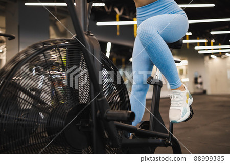 Cropped image woman in blue leggins pedaling on crossfit exercise bike Cropped image woman in blue leggins pedaling on crossfit exercise bike 88999385