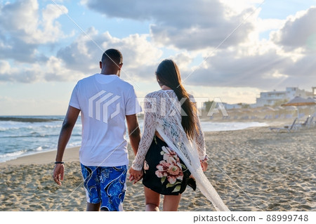 Young happy couple walking on the beach holding hands, back view. Young happy couple walking on the beach holding hands, back view. 88999748