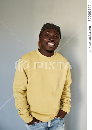 Portrait of smiling cool black student guy in cap and yellow sweater holding hands in jeans pockets against gray wall Portrait of smiling cool black student guy in cap and yellow sweater holding hands in jeans pockets against gray wall 89000163