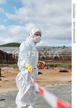 Pollution specialist in mask and protective suit marking off dangerous territory with red and white restrictive tape Pollution specialist in mask and protective suit marking off dangerous territory with red and white restrictive tape 89000563