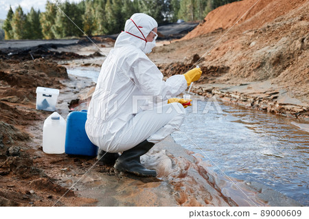 Land scientist in biohazmat suit, mask and gloves crouching at river and dropping water sample into container 89000609