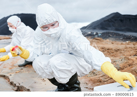Ecologist in respirator and safety goggles crouching on clay land and holding container of soil sample while working at toxic area Ecologist in respirator and safety goggles crouching on clay land and holding container of soil sample while working at toxic area 89000615