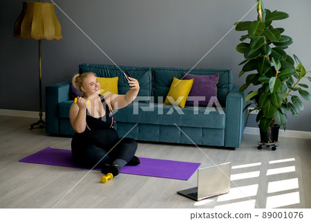 An obese young blonde woman with a dumbbell takes a selfie on her smartphone during a home workout. Sports at home in the living room with a green sofa and yellow-purple pillows. Model plus size An obese young blonde woman with a dumbbell takes a selfie on her smartphone during a home workout. Sports at home in the living room with a green sofa and yellow-purple pillows. Model plus size 89001076