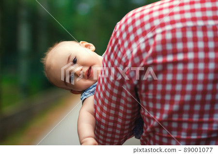 A father carries his little daughter in his arms. The child peeks out from behind Dad's broad shoulders. The child feels safe. The relationship between parents and children. Child psychology. A father carries his little daughter in his arms. The child peeks out from behind Dad's broad shoulders. The child feels safe. The relationship between parents and children. Child psychology. 89001077