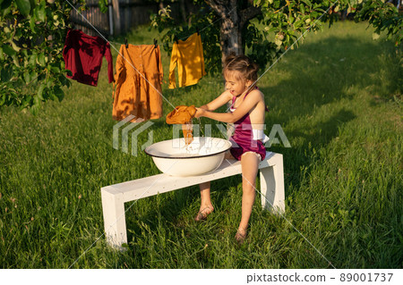 A little girl washes clothes with her hands in a basin, sitting on a bench in the garden. Laundry is dried on a clothesline under a tree.The child is happy to help his mother launder 89001737