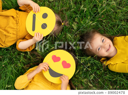 Three funny girlfriends are lying on the green grass with different emoticons in their hands. One girl is laughing, and two are holding smile masks covering their faces. Types of temperament. Three funny girlfriends are lying on the green grass with different emoticons in their hands. One girl is laughing, and two are holding smile masks covering their faces. Types of temperament. 89002478