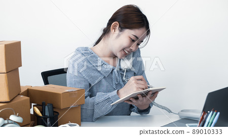 Young Asian woman business owner with many parcel boxes on the table happy online sales job, use the laptop, get an order from customers, take notes, and make arrangements for delivery by post 89010333