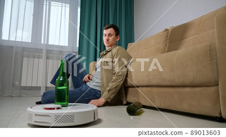 White robot vacuum cleaner delivers alcohol beer bottle to brunet man sitting on floor near sofa in living room 89013603