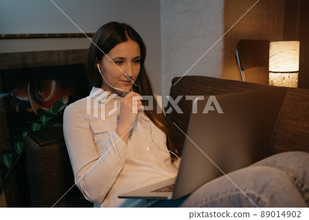A woman with headphones working remotely on her laptop sitting on the sofa 89014902
