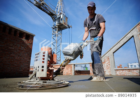 Specialist in arrangement of floor who leveling inequality of surface after installation of semi-dry screed by special machine. On the background tower crane and blue cloudless sky. Specialist in arrangement of floor who leveling inequality of surface after installation of semi-dry screed by special machine. On the background tower crane and blue cloudless sky. 89014935