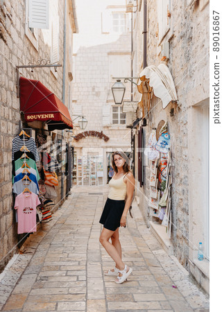Girl Tourist Walking Through Ancient Narrow Street On A Beautiful Summer Day In MEDITERRANEAN MEDIEVAL CITY, OLD TOWN BUDVA, MONTENEGRO. Young Beautiful Cheerful Woman Walking On Old Street At 89016867