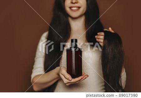Young woman with long healthy brown hair holding shampoo on palm, advertising hair-care products 89017390