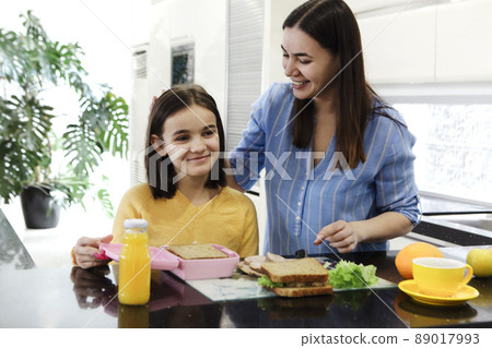 Cheerful ethnic mother making healthy lunch box with healthy food for her little daughter while cooking together at the white modern kitchen the morning time 89017993