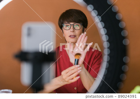 Middle-aged woman talking on cosmetics with makeup eye shadows and blush palette and brushes while recording her video. Mature female making video for her blog on cosmetics Middle-aged woman talking on cosmetics with makeup eye shadows and blush palette and brushes while recording her video. Mature female making video for her blog on cosmetics 89024391