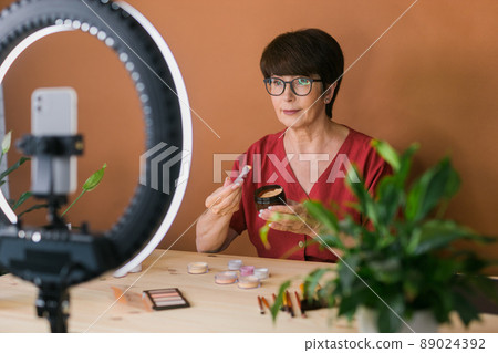 Middle-aged woman talking on cosmetics with makeup eye shadows and blush palette and brushes while recording her video. Mature female making video for her blog on cosmetics 89024392