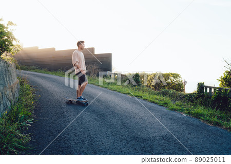 Pensive young male in casual wear standing on asphalt road dreaming about something on sunset 89025011