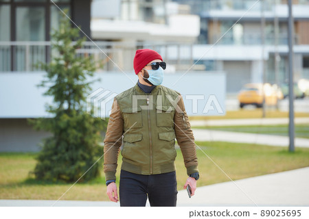 A man in a medical face mask holding his smartphone and walking down the street. 89025695