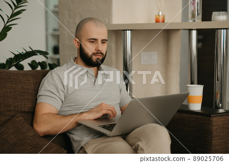 A bald man with a beard works remotely on his laptop sitting on a sofa at home. 89025706