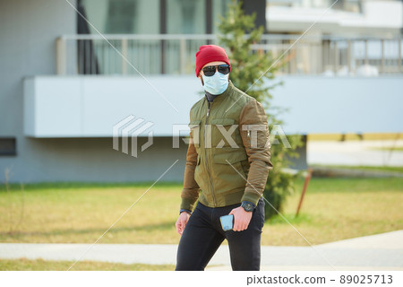 A man in a medical face mask holding his smartphone and walking down the street. A man in a medical face mask holding his smartphone and walking down the street. 89025713