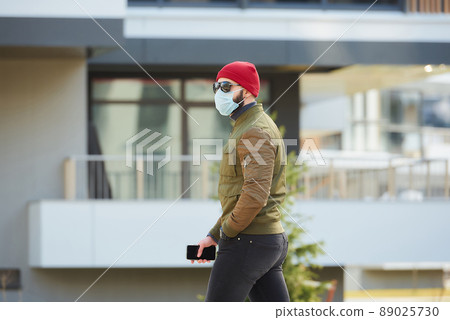 A man in a medical face mask holding his smartphone and walking down the street. A man in a medical face mask holding his smartphone and walking down the street. 89025730