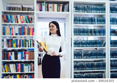 Confident Asian woman with books and disposable cup of coffee looking away 89026305
