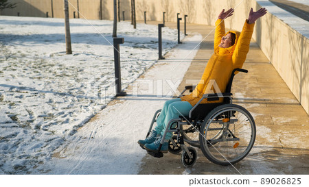 Happy woman in wheelchair happily pulls her hands up while walking in winter park.  89026825