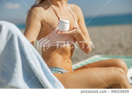 Woman in swimsuit holding, advertising jar with skin care product while relaxing on beach 89028809