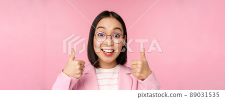 Awesome, congrats. Face of excited asian businesswoman in glasses, smiling pleased, showing thumbs up in approval, standing over pink background 89031535