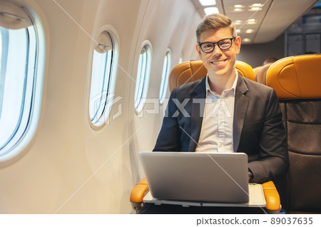 During a flight, a businessman in formal attire and glasses looks out the plane window while working on a laptop computer. Service with Internet access on board and a business travel concept During a flight, a businessman in formal attire and glasses looks out the plane window while working on a laptop computer. Service with Internet access on board and a business travel concept 89037635