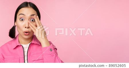 Close up portrait of young asian girl looking surprised, express amazement and wonder, peeking through fingers, standing over pink background 89052605